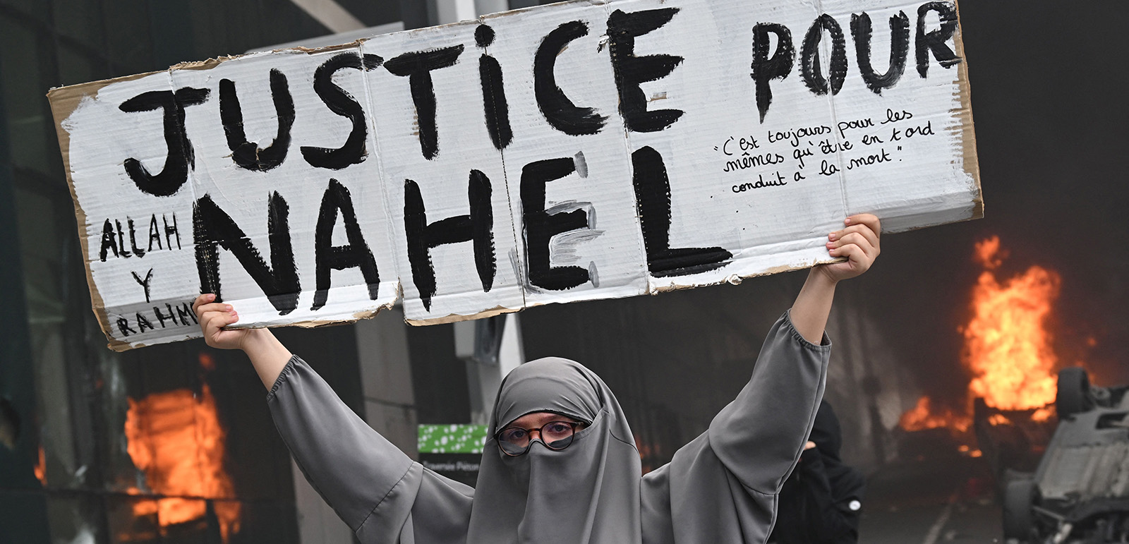 An attendee holds a banner reading "Justice for Nahel" as cars burn in the street at the end of a commemoration march for a teenage driver shot dead by a policeman, in the Parisian suburb of Nanterre, on June 29, 2023. Violent protests broke out in France in the early hours of June 29, 2023, as anger grows over the police killing of a teenager, with security forces arresting 150 people in the chaos that saw balaclava-clad protesters burning cars and setting off fireworks. Nahel M., 17, was shot in the chest at point-blank range in Nanterre in the morning of June 27, 2023, in an incident that has reignited debate in France about police tactics long criticised by rights groups over the treatment of people in low-income suburbs, particularly ethnic minorities. (Photo by Bertrand GUAY / AFP) (Photo by BERTRAND GUAY/AFP via Getty Images)