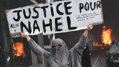 An attendee holds a banner reading "Justice for Nahel" as cars burn in the street at the end of a commemoration march for a teenage driver shot dead by a policeman, in the Parisian suburb of Nanterre, on June 29, 2023. Violent protests broke out in France in the early hours of June 29, 2023, as anger grows over the police killing of a teenager, with security forces arresting 150 people in the chaos that saw balaclava-clad protesters burning cars and setting off fireworks. Nahel M., 17, was shot in the chest at point-blank range in Nanterre in the morning of June 27, 2023, in an incident that has reignited debate in France about police tactics long criticised by rights groups over the treatment of people in low-income suburbs, particularly ethnic minorities. (Photo by Bertrand GUAY / AFP) (Photo by BERTRAND GUAY/AFP via Getty Images)