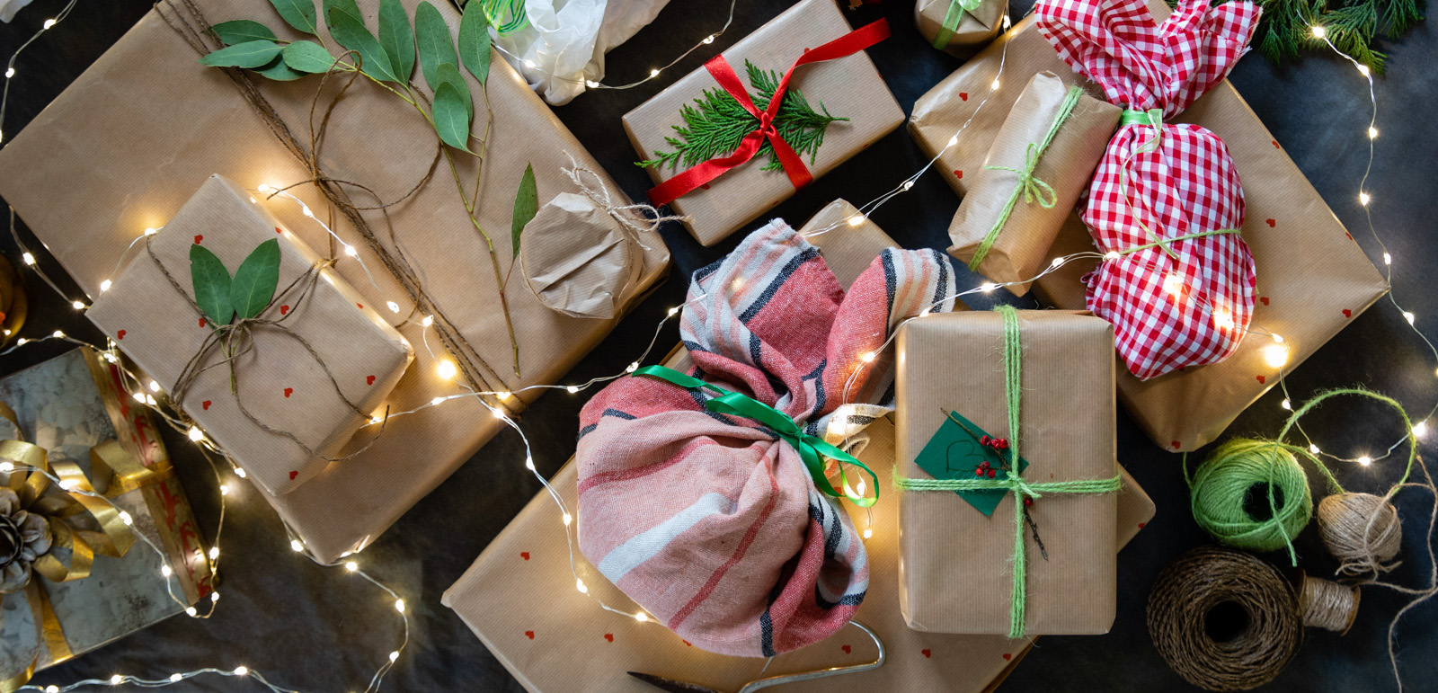 An overhead view of wrapped Christmas presents on a backdrop which have been decorated with ribbon, string, eucalyptus leaves and holly leaves. They are ready to be given at Christmas time and they are on the table, draped in Christmas lights.
