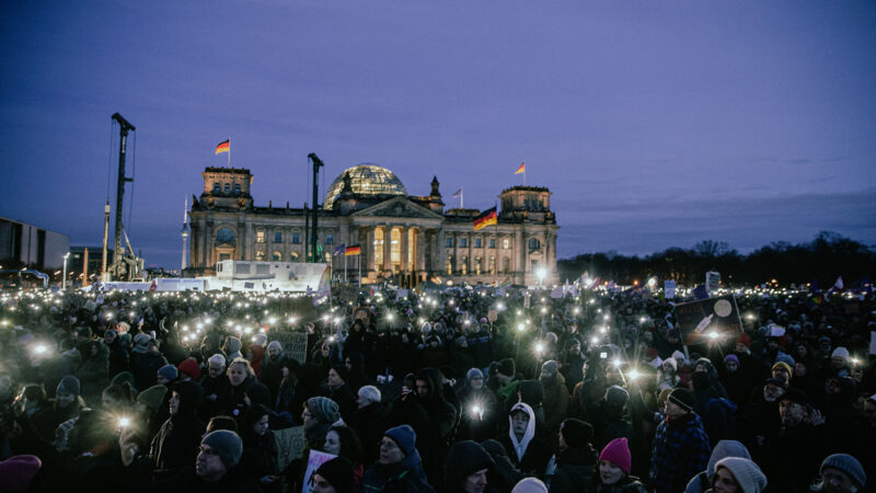 A crowd of people assembled in front of the Bundestag building in Berlin. People use their phones for light in the evening.