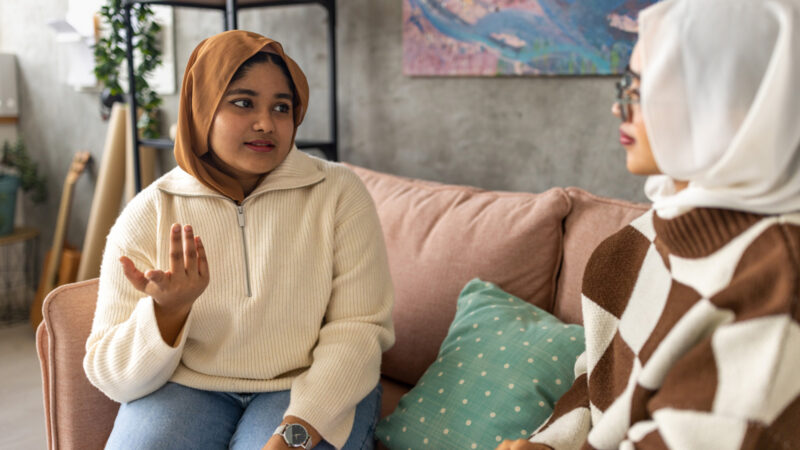 Two young women talking to each other while sitting on sofa at home.