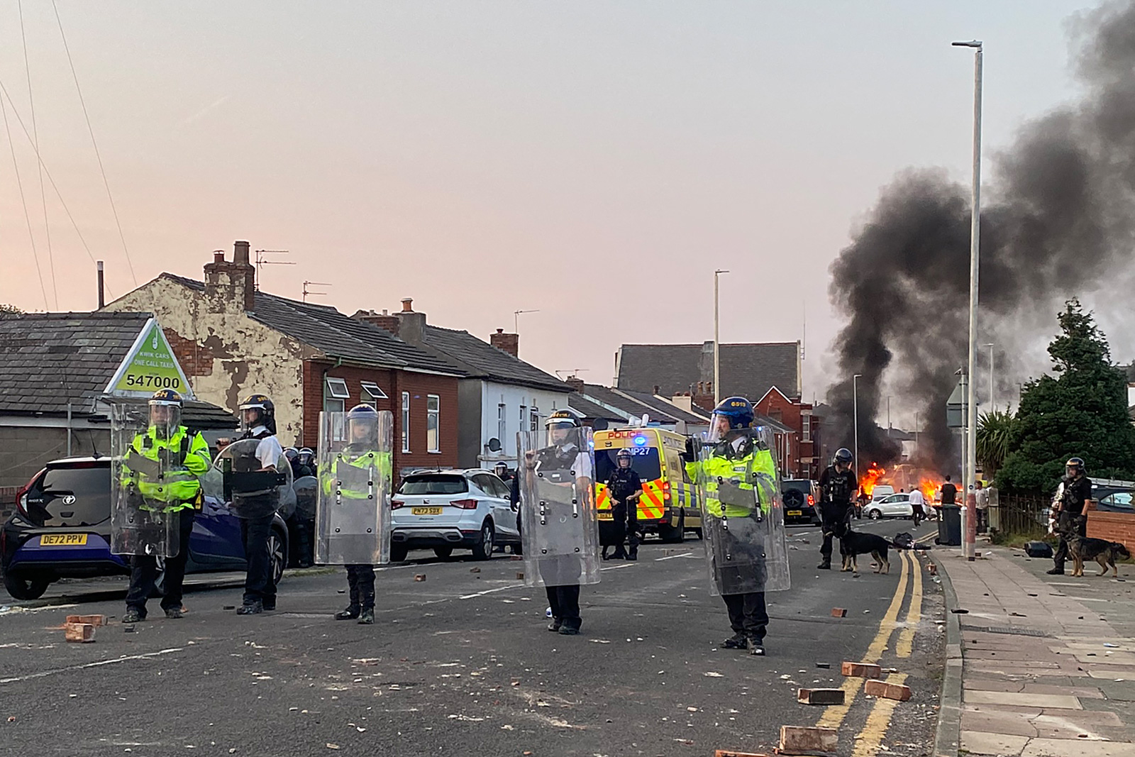 Smoke billows from a fire started by protesters as riot police stand guard after disturbances near the Southport Islamic Society Mosque in Southport, northwest England, on July 30, 2024, a day after a deadly child knife attack. Violent clashes broke out in the northern England town where a knife attack claimed the lives of three children, with around 100 protesters lighting fires and battling police. A 17-year-old male suspect from a nearby village arrested shortly after the incident remained in custody, police added, as they warned against speculating about his identity or details of the investigation. Photo by Roland LLOYD PARRY / AFP