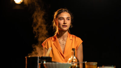 A young woman wearing an orange shirt cooks a Persian dish while telling a story from her life in the play My English Persian Kitchen
