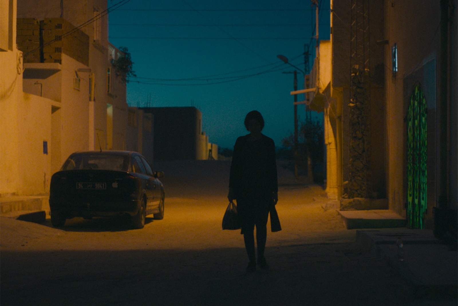 Film still from Aïcha, 2024, shows a woman walking down an abandoned village road at night