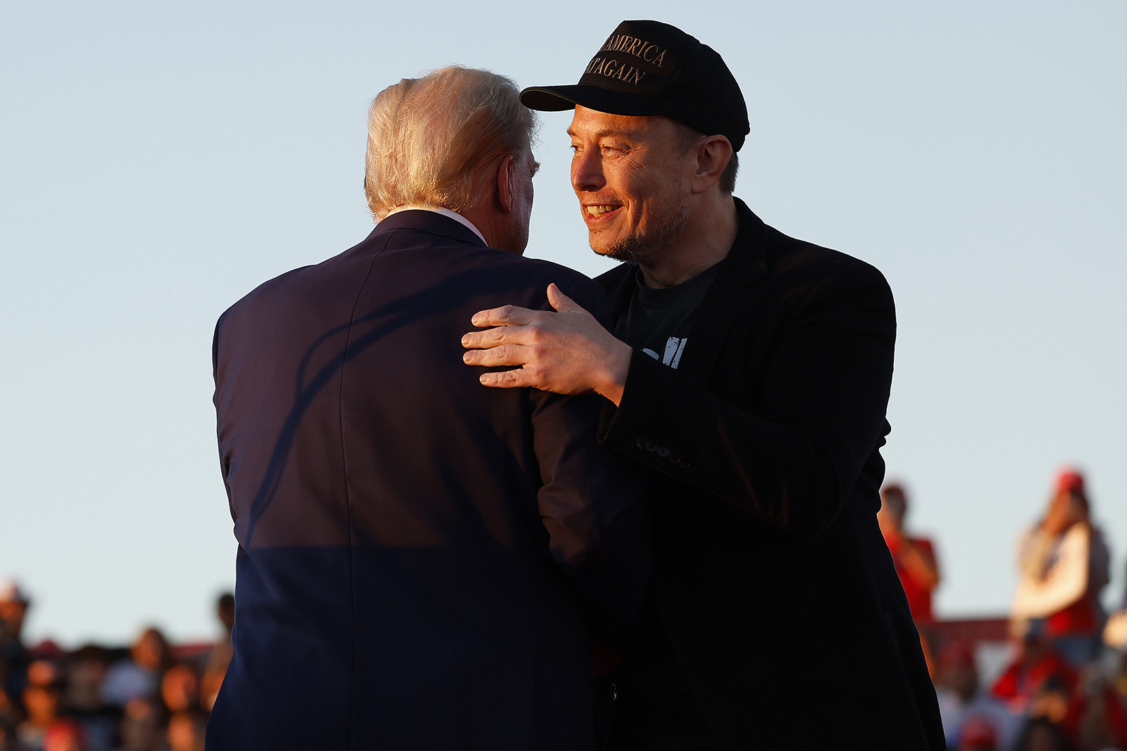 Elon Musk, wearing a Make America Great Again cap and facing the camera, hugs Donald Trump, who faces away from the camera