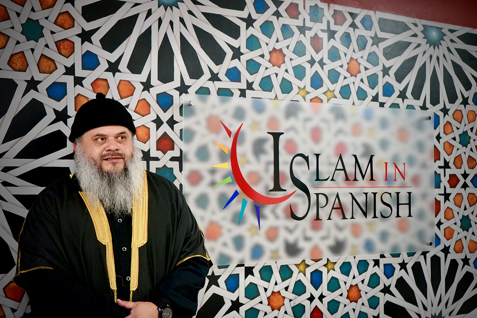 Sheikh Omar Hernandez standing in front of a colourful, patterned sign for IslamInSpanish