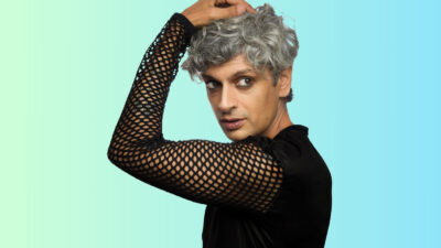 Kazim Ali wearing a black mesh top with one arm raised and a hand on his grey hair