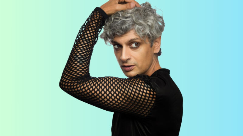 Kazim Ali wearing a black mesh top with one arm raised and a hand on his grey hair