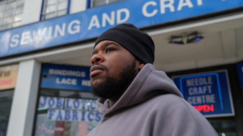 Ridwan Fadlu-Deen wears a grey hoodie and black beanie and stands in front of a blue and white building
