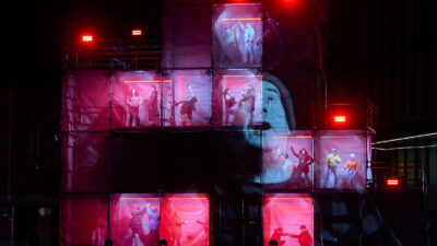 Performers inside giant boxes stacked on each other, with a face projected on the outside of the structure