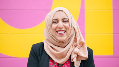Akeela Ahmed, smiling and dressed in a black jacket and a pink hijab, against a pink and yellow background