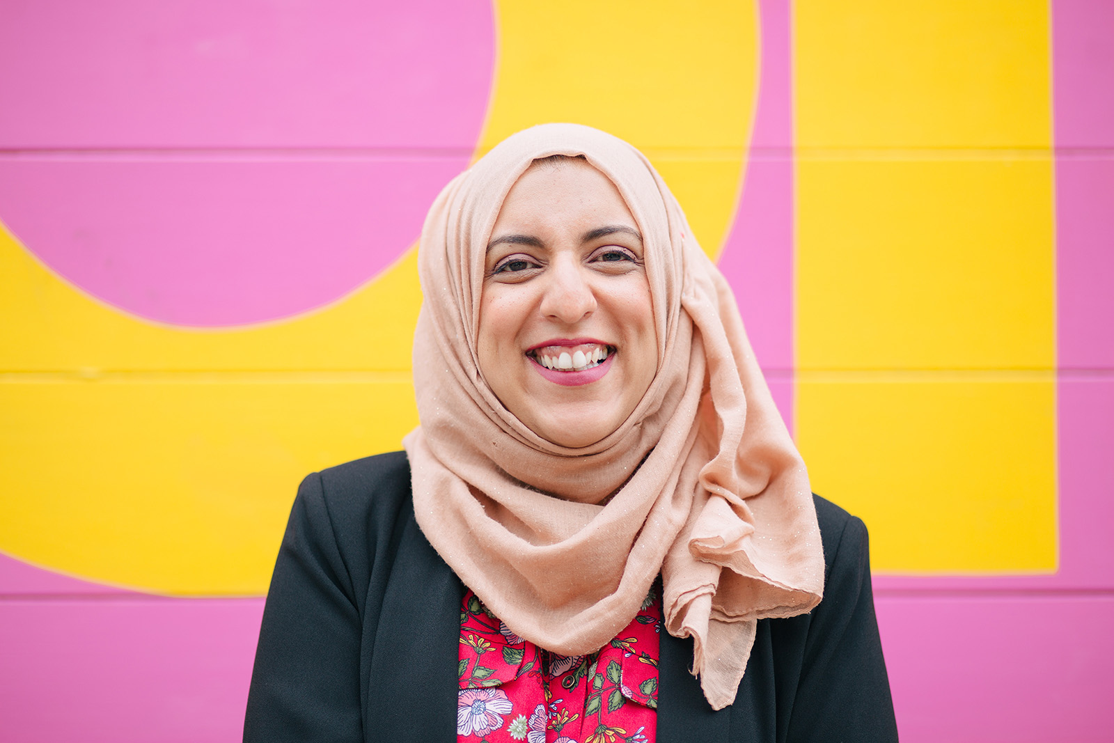 Akeela Ahmed, smiling and dressed in a black jacket and a pink hijab, against a pink and yellow background