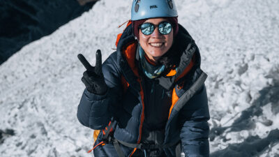 Freshta Ibrahimi on a snowy mountain wearing climbing gear, a helmet and sunglasses and giving the V sign