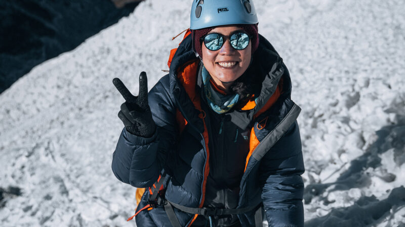Freshta Ibrahimi on a snowy mountain wearing climbing gear, a helmet and sunglasses and giving the V sign