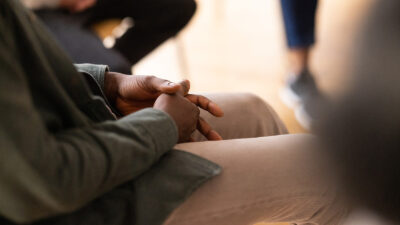 Close-up of a man sitting in a circle during group therapy with his hands clasped. Cropped shot of participants in mental health therapy session.