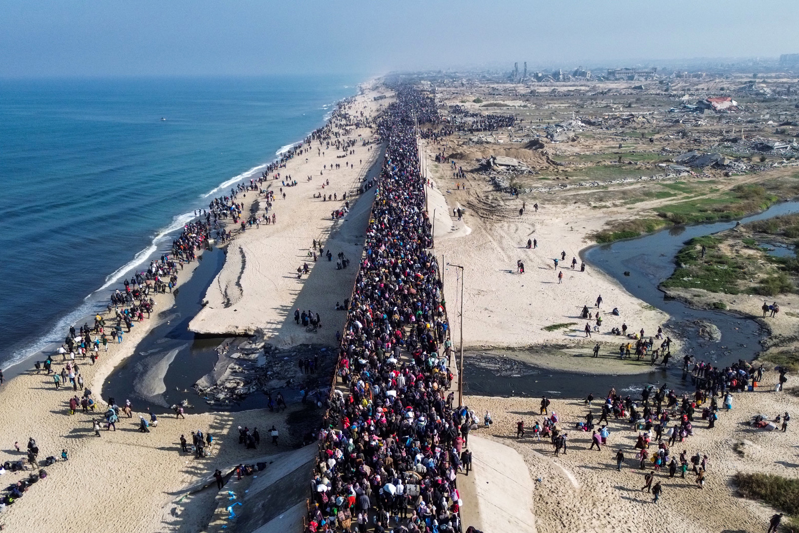 An aerial picture of a road filled with people flanked by the coast on the left and a rubble-strewn landscape on the right