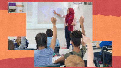 A teacher pointing to a whiteboard in a classroom, in front of pupils holding up their hands