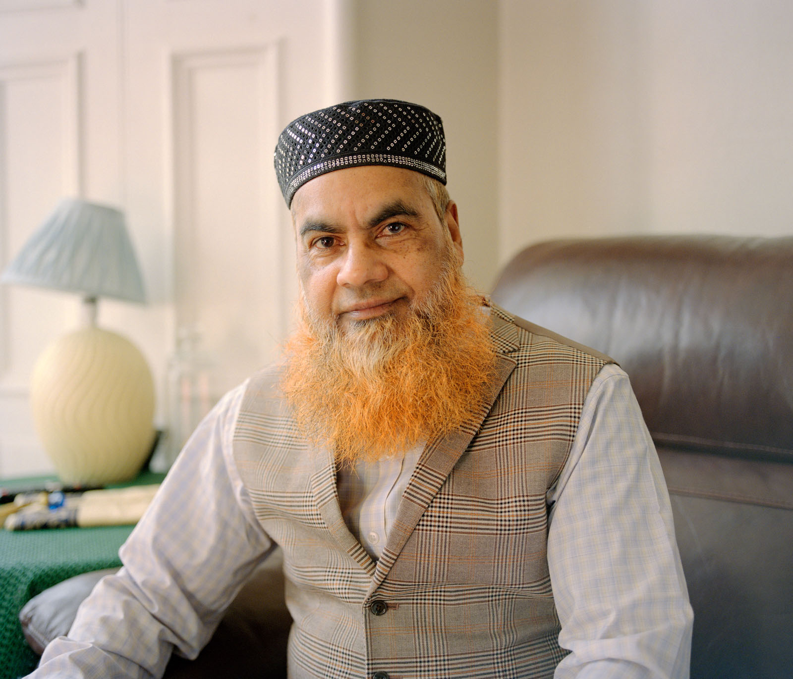 An older British Pakistani Muslim man sitting on a brown leather sofa.