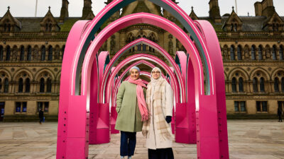 Two figures in green and cream stand beneath a structure of multiple arches constructed in hot pink
