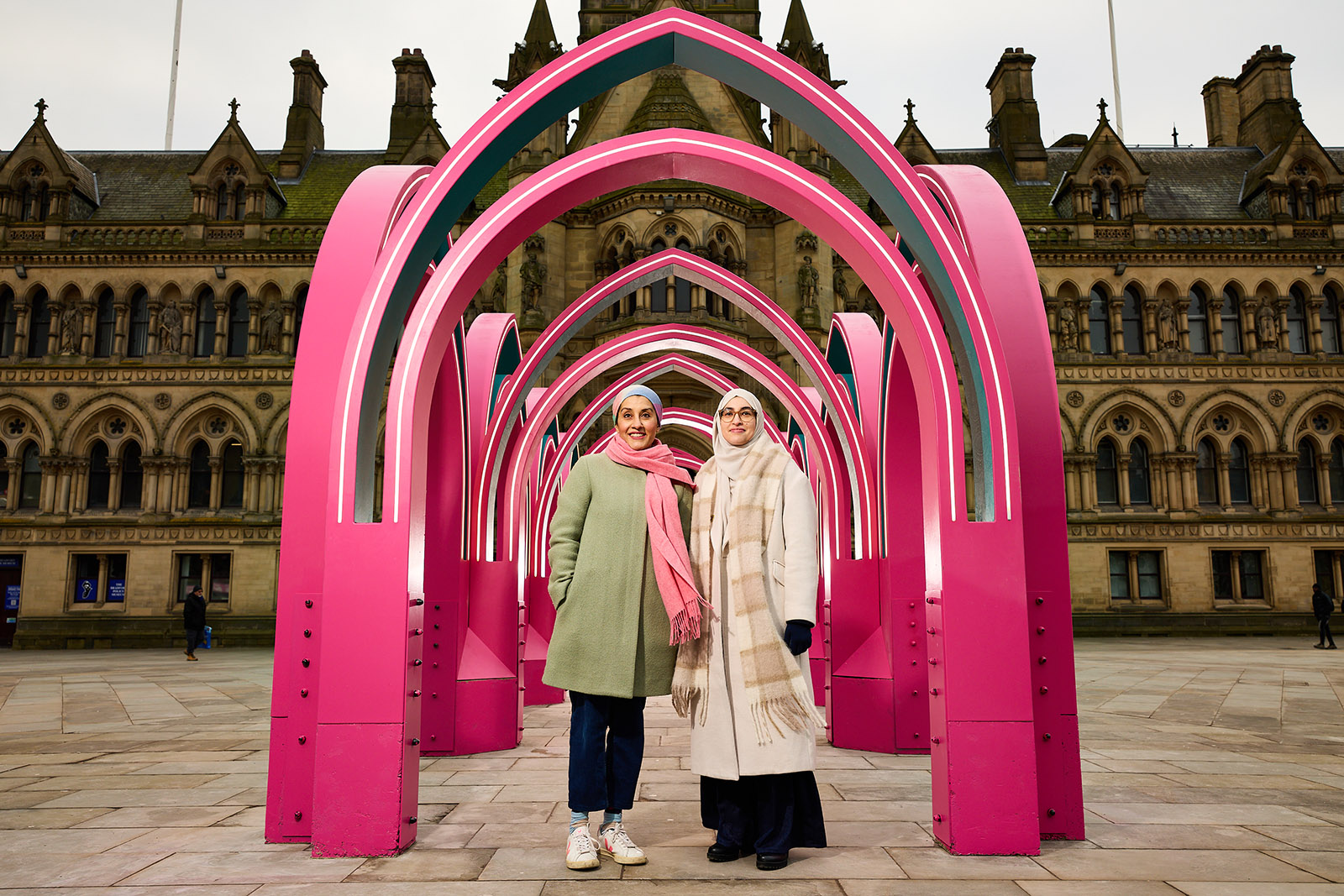 Two figures in green and cream stand beneath a structure of multiple arches constructed in hot pink