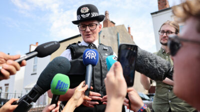 A police officer in uniform surrounded by outstretched arms with microphones