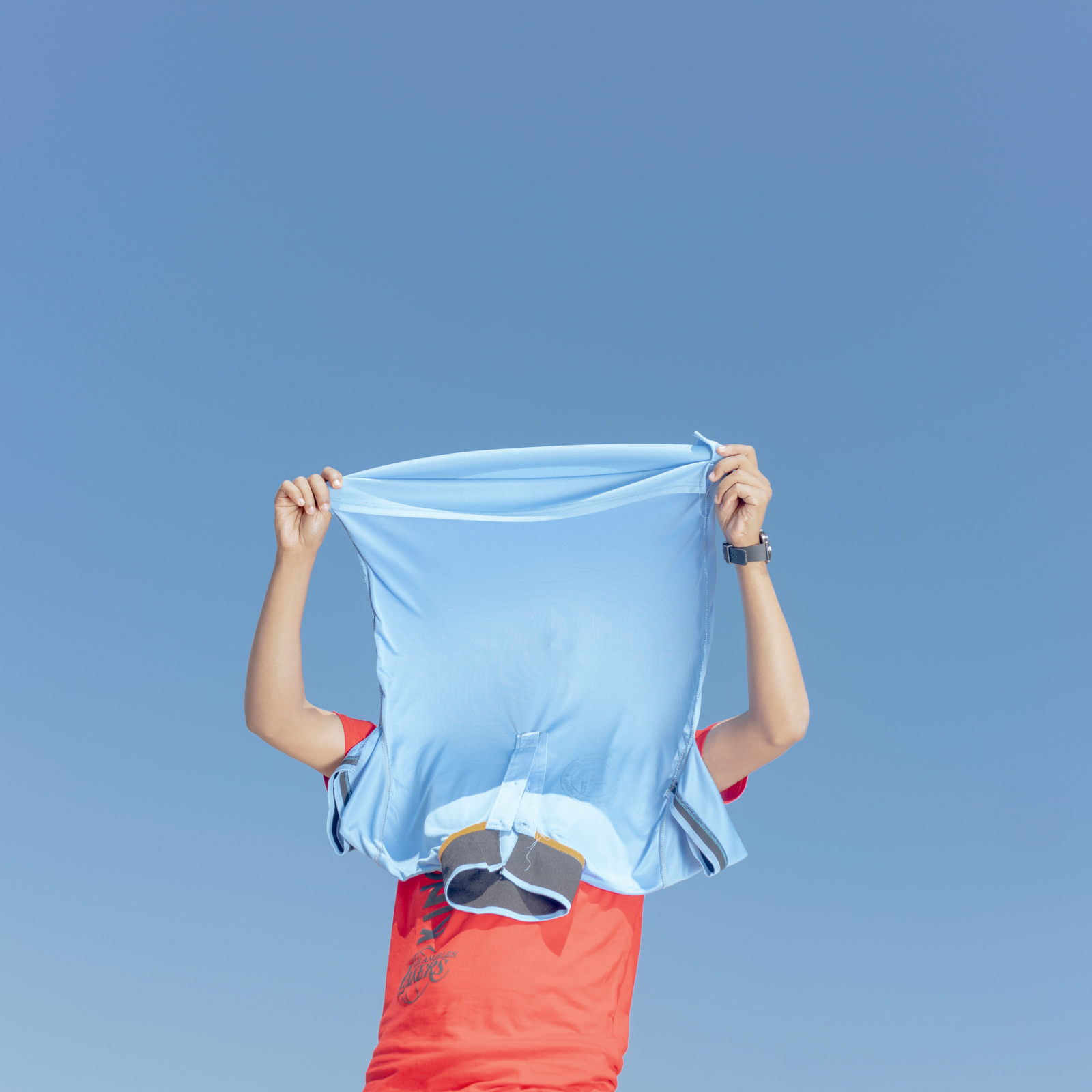 A young boy holds a blue T-shirt over his head against a blue sky