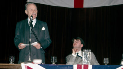 Anthony Murphy sits on stage while Enoch Powell speaks at an event in 1989