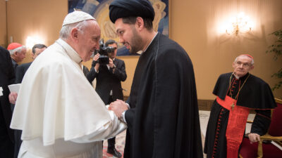 The pope, in a white robe and cap, shakes hands with Sayed Ali Abbas Razawi, dressed in black. The two men are turned towards each other with their expressions warm and their faces close