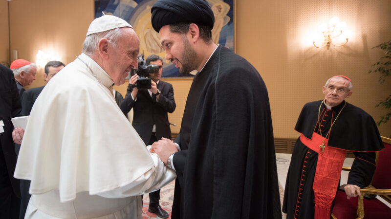 The pope, in a white robe and cap, shakes hands with Sayed Ali Abbas Razawi, dressed in black. The two men are turned towards each other with their expressions warm and their faces close