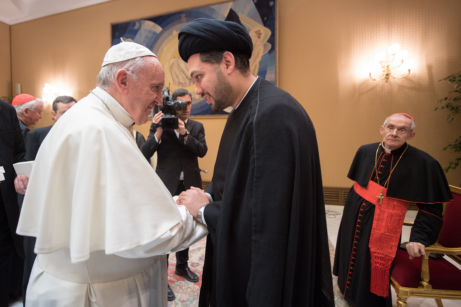 The pope, in a white robe and cap, shakes hands with Sayed Ali Abbas Razawi, dressed in black. The two men are turned towards each other with their expressions warm and their faces close