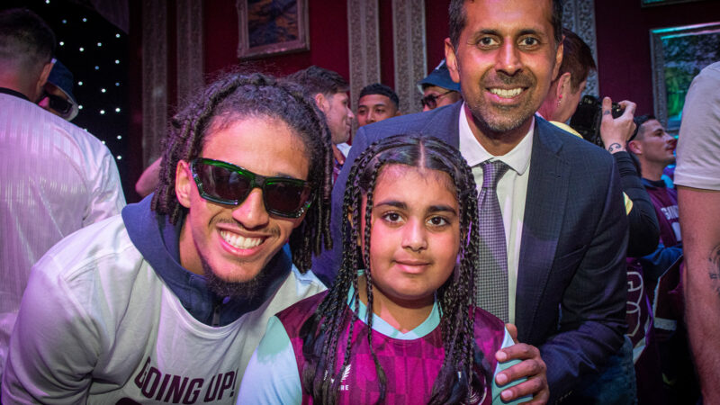 A man in sunglasses and an older man in a suit smile as they stand either side of a little girl in a purple jumper, also smiling