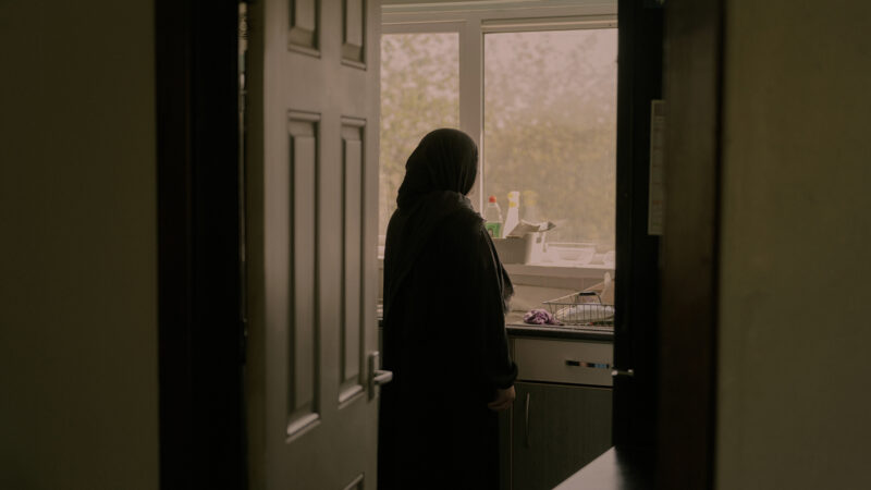 A woman in hijab is silhouetted against a kitchen window. The light is dim and she gazes out the window. The picture is taken through an open door.