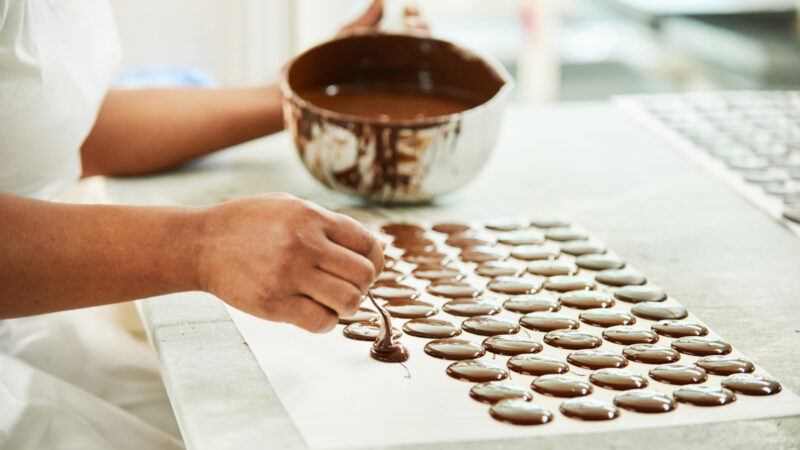 Close-up of a worker in a chocolate factory pouring circles of melted chocolate onto a baking sheet to cool