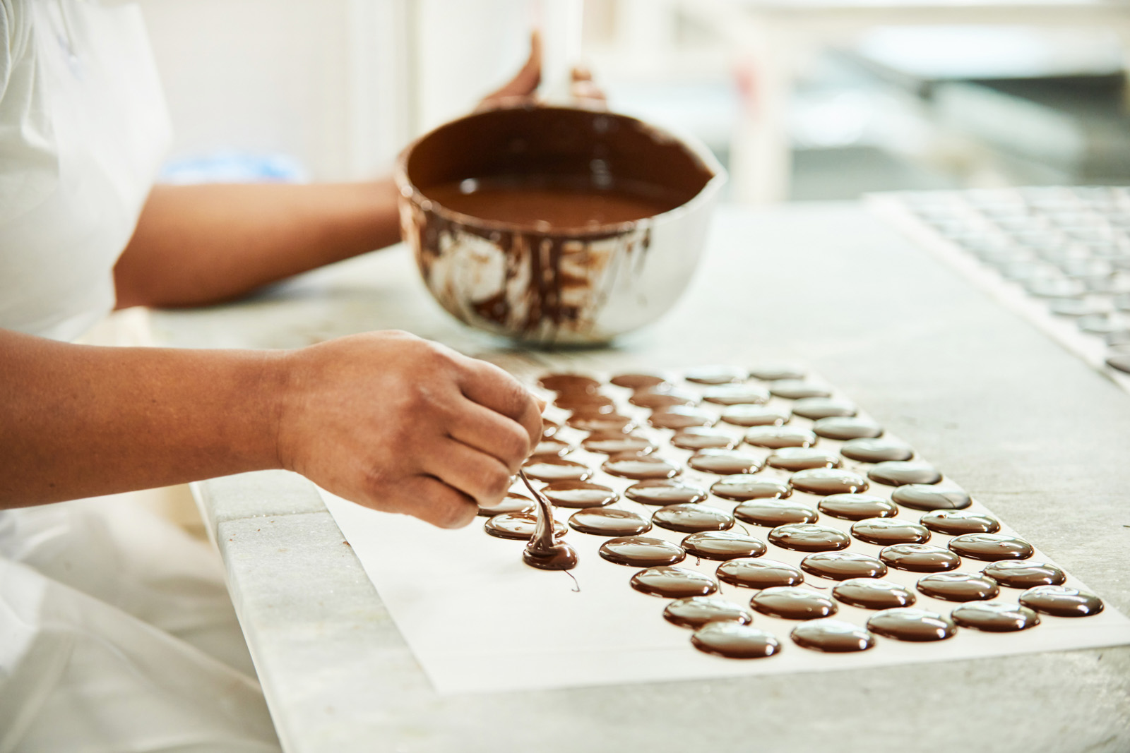Close-up of a worker in a chocolate factory pouring circles of melted chocolate onto a baking sheet to cool