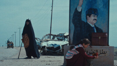 A still from The President's Cake showing a woman in an abaya standing by a road