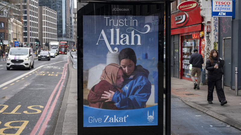 A bus station poster advertising zakat giving shows two Muslim Women embracing and text that reads 'Trust in Allah'.