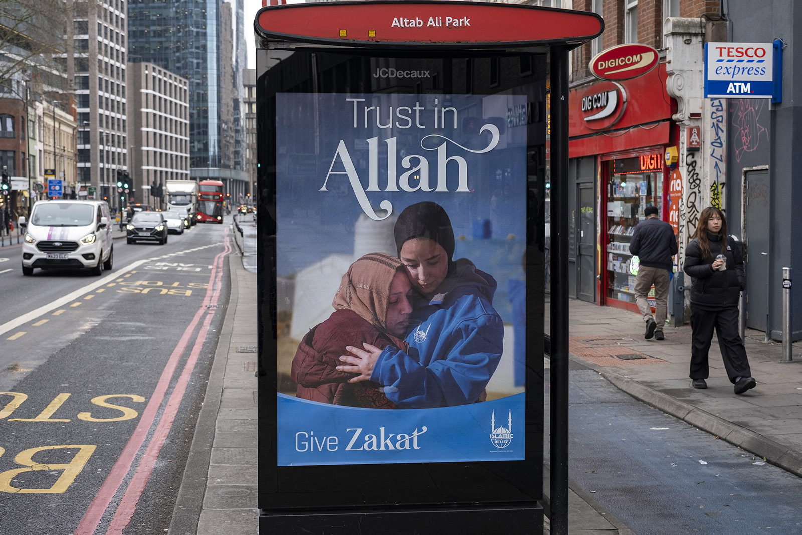 A bus station poster advertising zakat giving shows two Muslim Women embracing and text that reads 'Trust in Allah'.