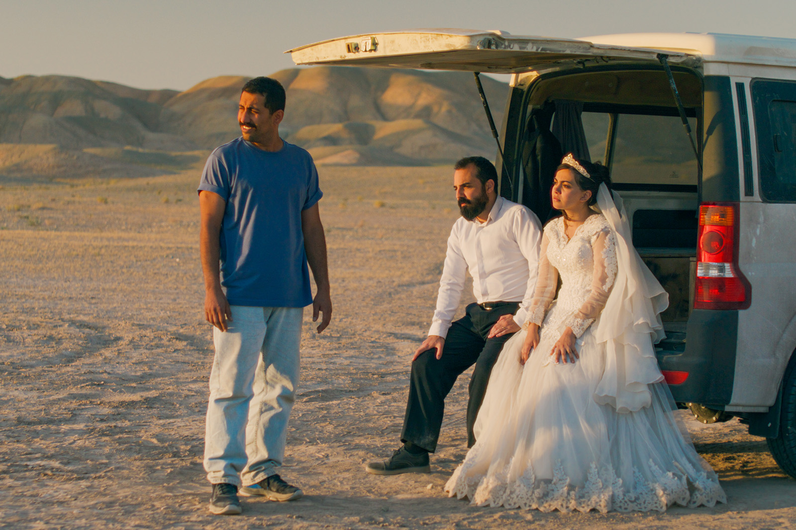 A bride and groom sit in the back of an open van in the desert