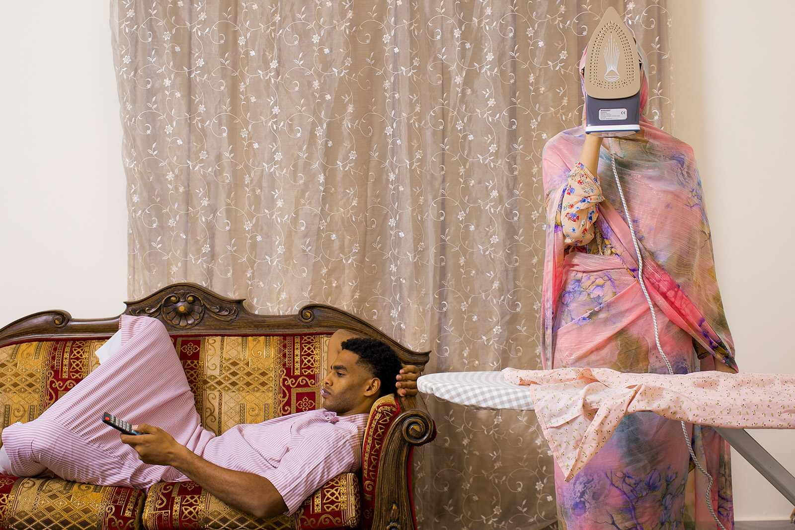 A Muslim woman wearing an elaborate dress irons while a man lies on a sofa with a TV remote in his hand