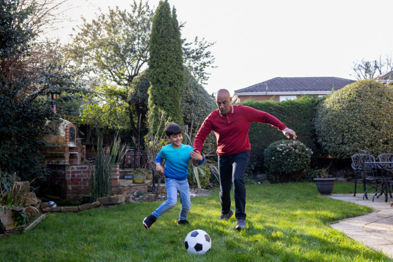 Full shot of a grandfather and grandson playing football with each other in the family garden with the sun shining behind them. They are both wearing casual clothing and sitting in a conservatory. Located in Middlesborough, England.