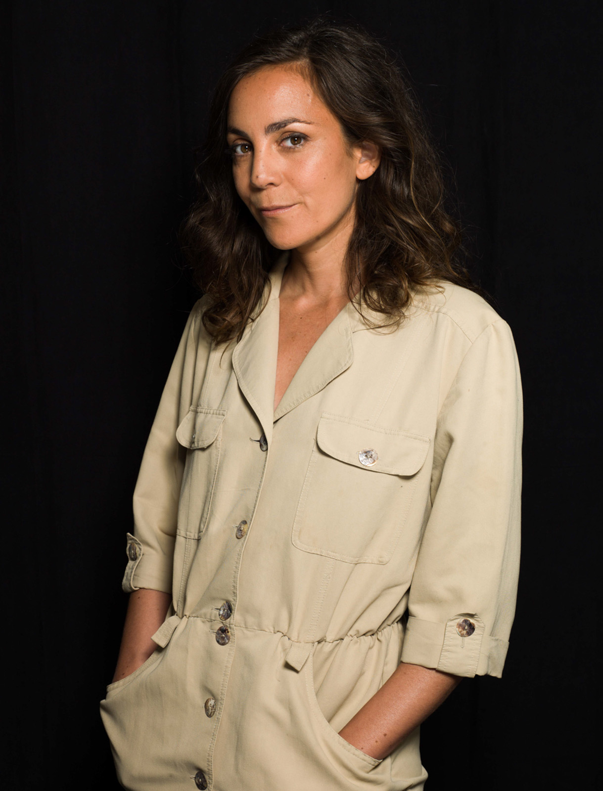 Hind Meddeb wearing a cream-coloured jumpsuit with her hands in her pockets, against a black background