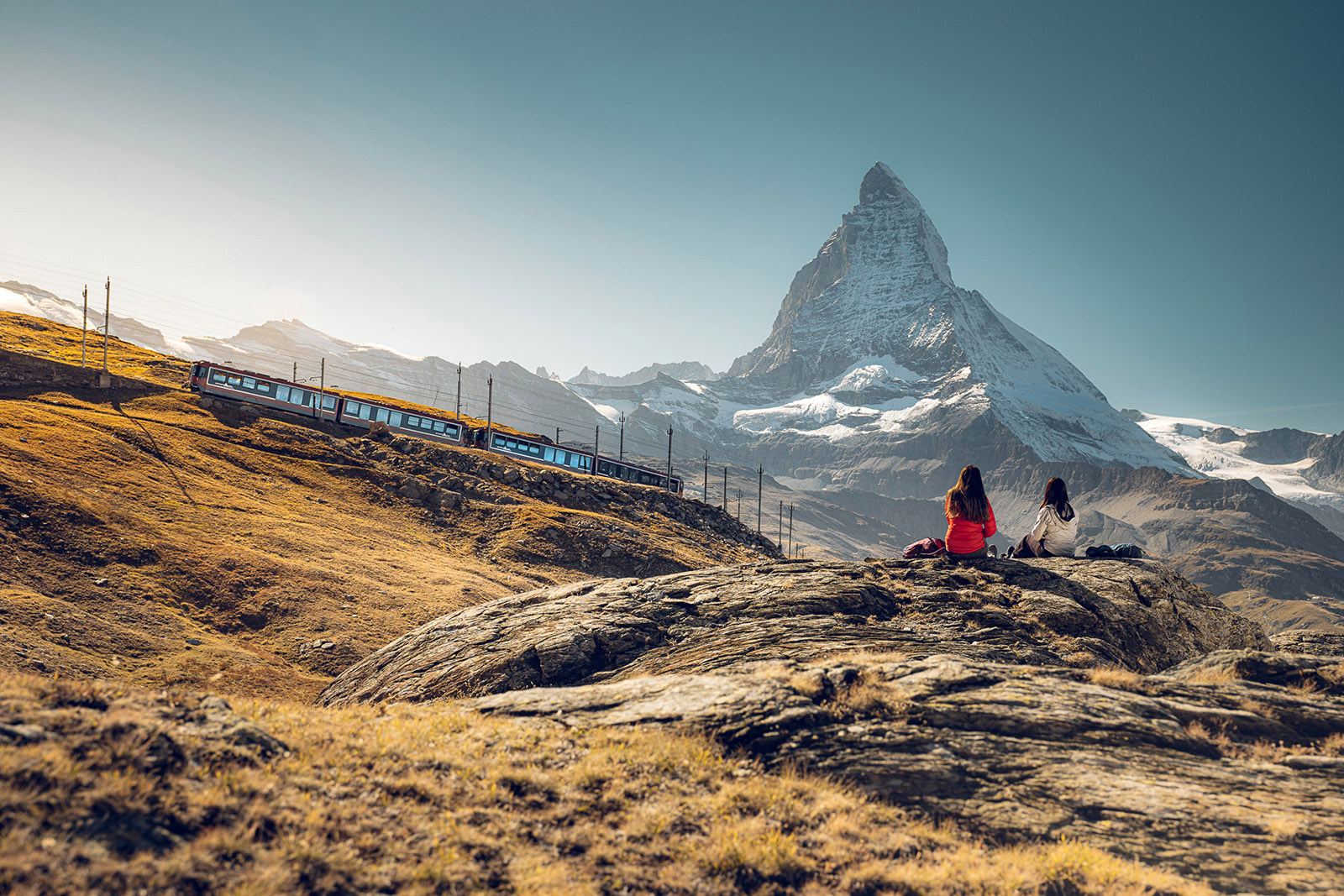 Two hikers watch a GGB mountain rack railway train pass by, with the Matterhorn in the background.