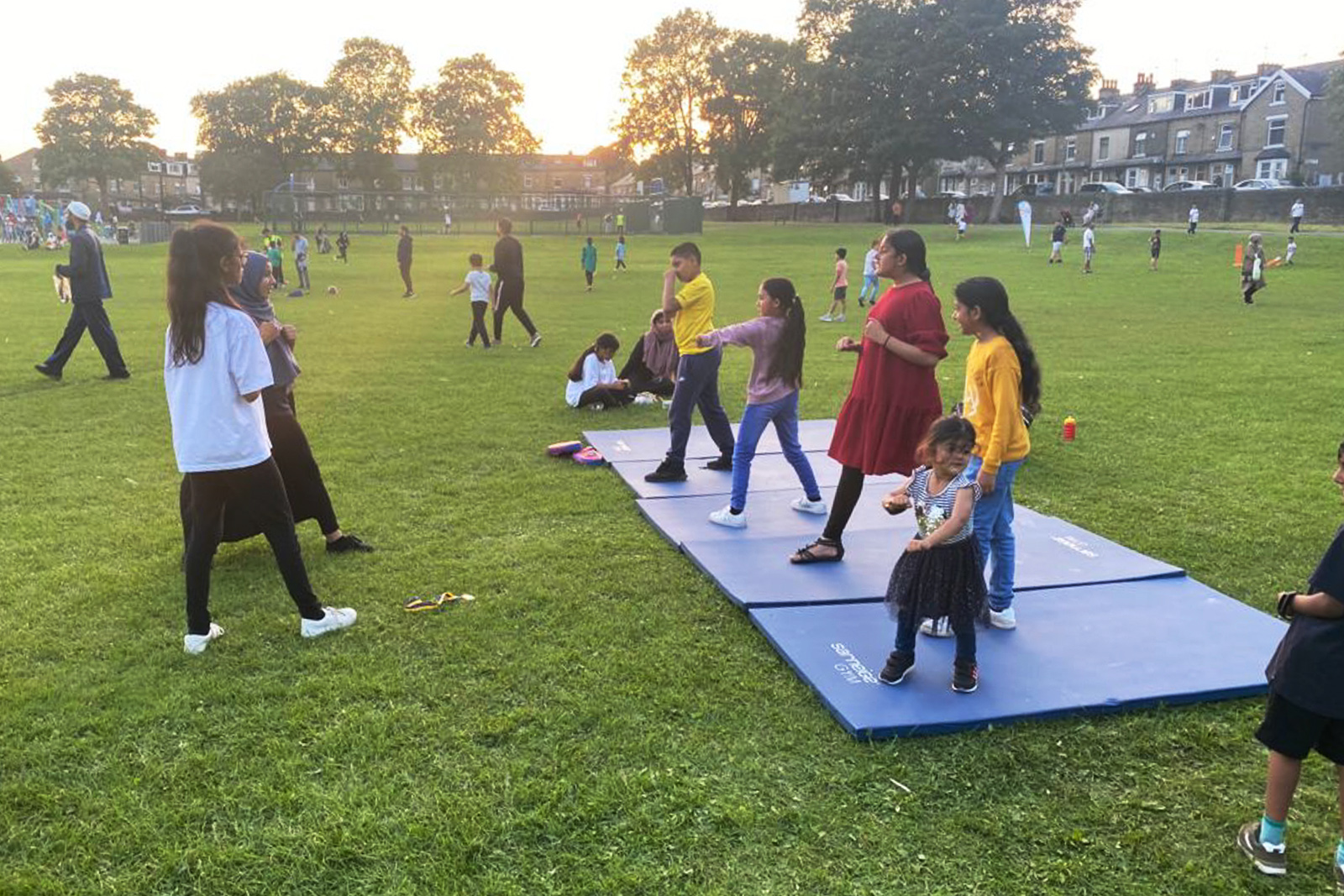 Girls strike martial arts posts on a row of mats in a park, facing a teacher