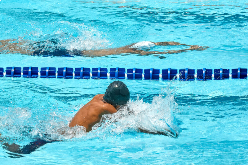 A swimmer in a blue cap, only their back and part of their head visible, swims in a lane with a blue divider behind them. A new generation of Muslim swimming teachers is being trained up by Newport charity KidCare4U