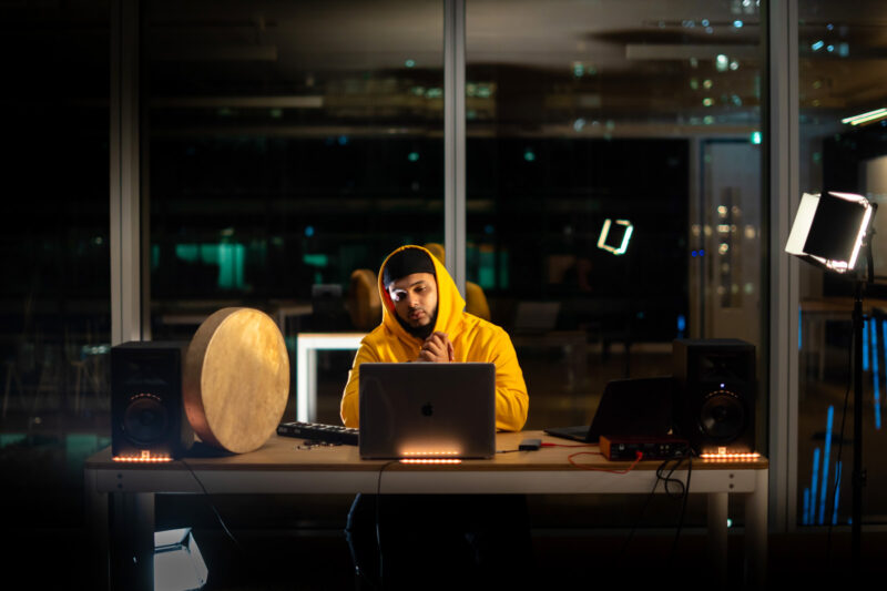 Photograph of producer Halal Beats, real name Umar Value, wearing a yellow hoodie in a recording studio behind a laptop.