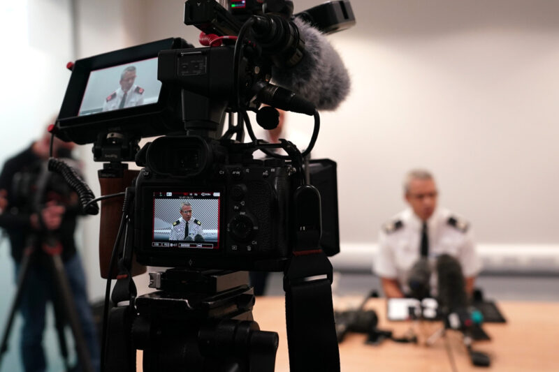 Video cameras focus on a police officer in uniform, seated at a desk as he addresses the room.