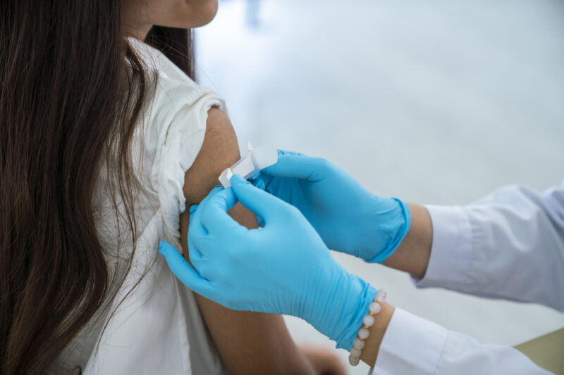 A close-up of a person's hands, in blue gloves, putting a plaster over someone's arm after a childhood vaccination