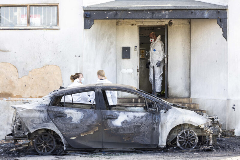 A burnt out car in front of a doorway with one door open. A person in a white forensic suit is in the doorway.