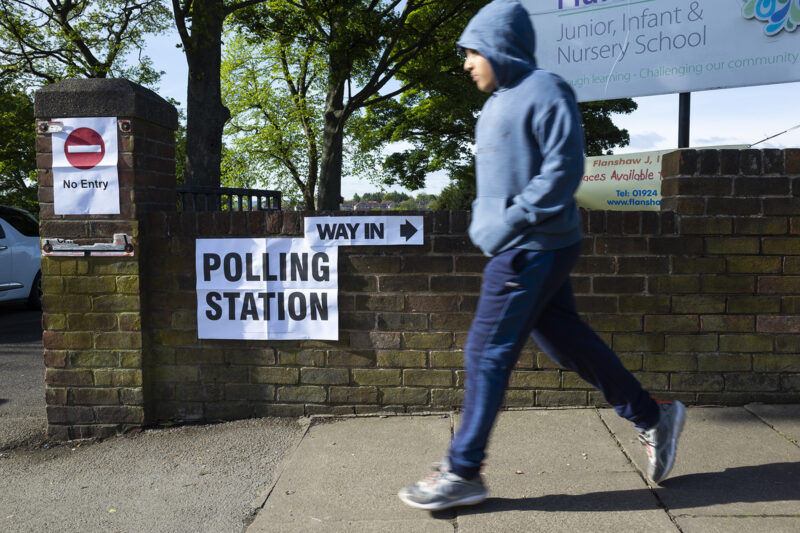 A small child, blurred in motion, runs past a sign that uses arrows to point the way towards the nearest local polling station, illustrating a story about votes at 16 and the impact on the size of the UK's Muslim electorate