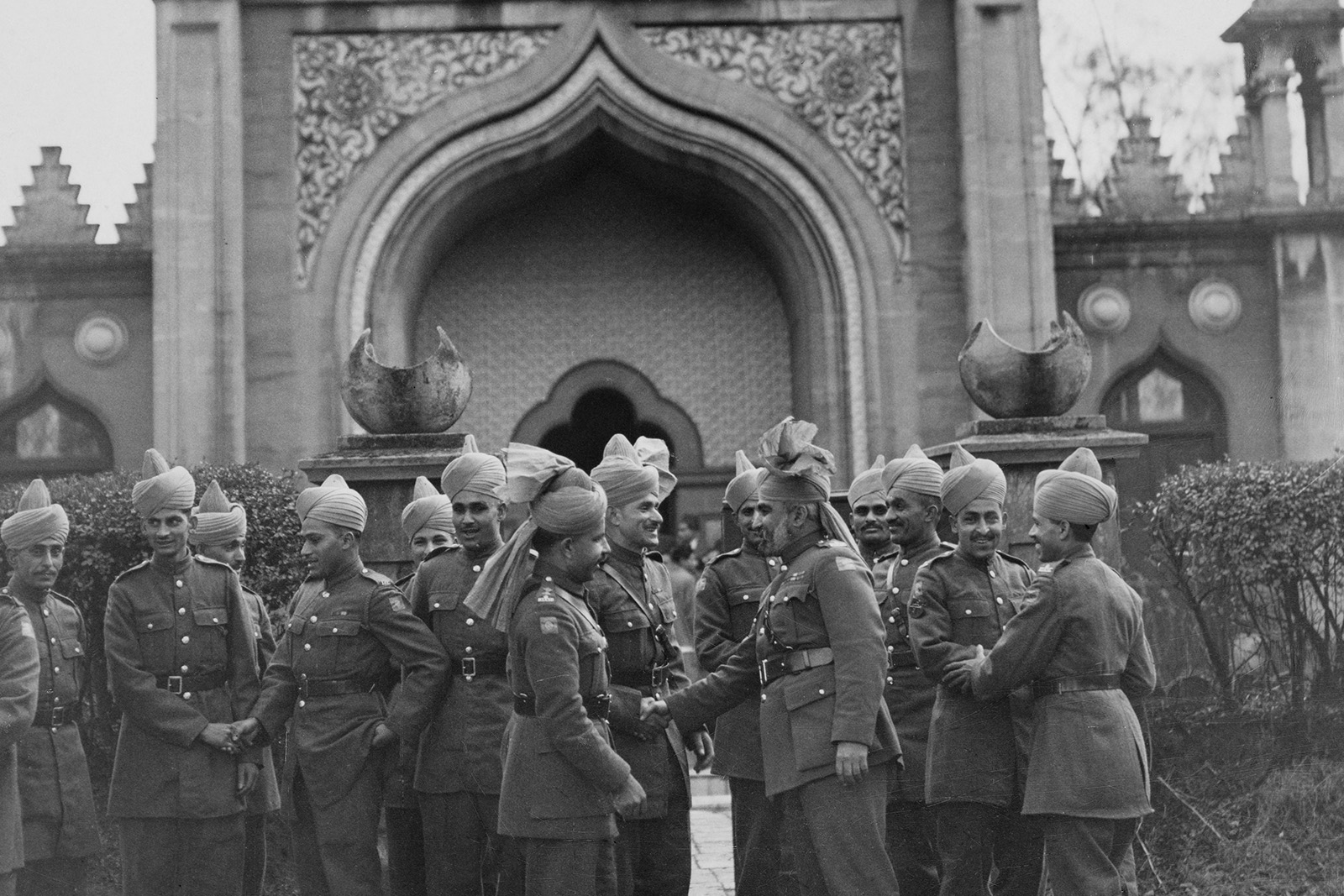 A black and white photo of men in military uniform and turbans, smiling and shaking hands, outside a mosque.